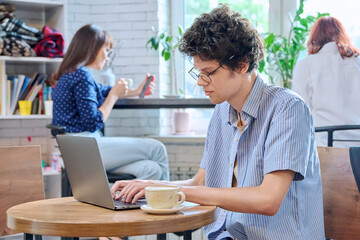 Guy college student typing on laptop, sitting at table in coffee shop with cup of coffee.