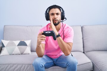 Hispanic young man playing video game holding controller sitting on the sofa hand on mouth telling...
