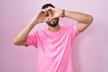 Hispanic young man standing over pink background doing heart shape with hand and fingers smiling...