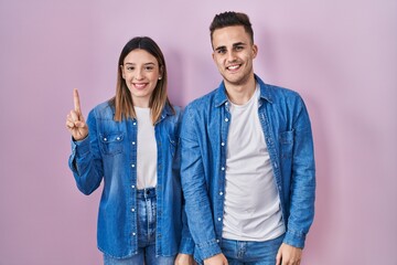 Young hispanic couple standing over pink background showing and pointing up with finger number one while smiling confident and happy.