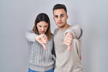 Young hispanic couple standing over white background looking unhappy and angry showing rejection and negative with thumbs down gesture. bad expression.