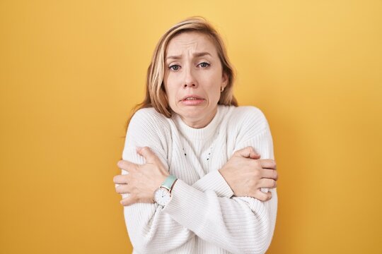 Young caucasian woman wearing white sweater over yellow background shaking and freezing for winter cold with sad and shock expression on face