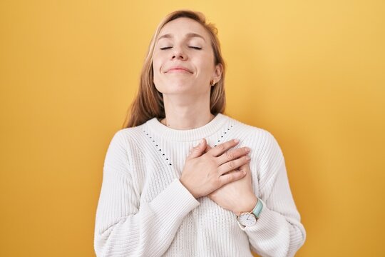 Young caucasian woman wearing white sweater over yellow background smiling with hands on chest with closed eyes and grateful gesture on face. health concept.