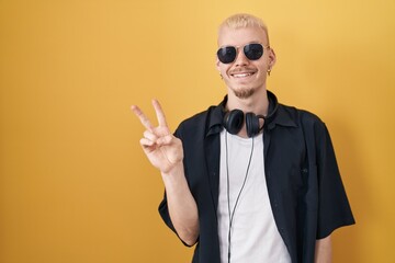 Young caucasian man wearing sunglasses standing over yellow background smiling with happy face winking at the camera doing victory sign with fingers. number two.