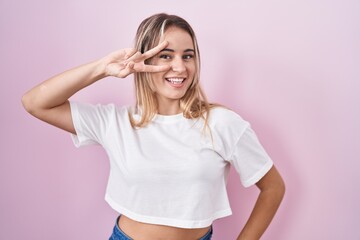 Young blonde woman standing over pink background doing peace symbol with fingers over face, smiling cheerful showing victory