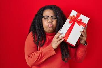 Plus size hispanic woman holding gift puffing cheeks with funny face. mouth inflated with air, catching air.