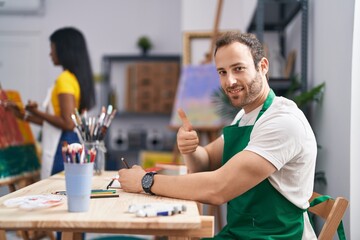 Hispanic man at painter studio smiling happy and positive, thumb up doing excellent and approval sign