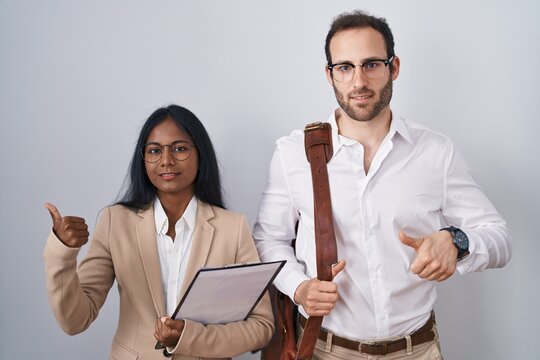 Interracial business couple wearing glasses pointing to the back behind with hand and thumbs up, smiling confident
