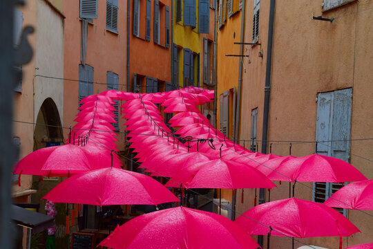 
Brightly colored pink umbrella's in a row over central shopping street in downtown Grasse, France, Europe, during early evening lit street, against multi-colored walls or facades - rainy day, wet umb