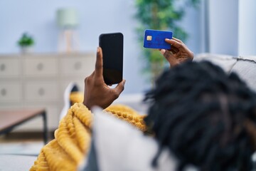 African american man using smartphone and credit card lying on sofa at home