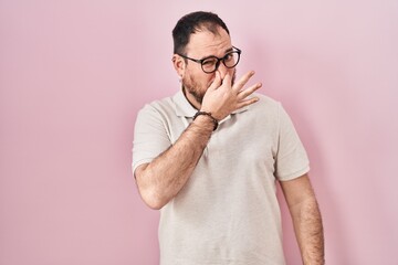 Plus size hispanic man with beard standing over pink background smelling something stinky and...