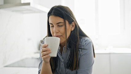 Young beautiful hispanic woman drinking a coffee leaning on the counter at the kitchen
