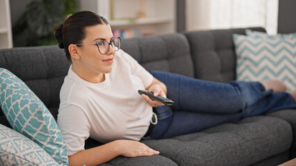 Young beautiful hispanic woman watching tv lying on sofa at home