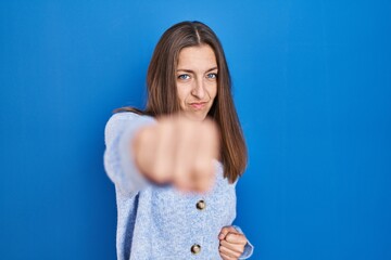 Young woman standing over blue background punching fist to fight, aggressive and angry attack,...