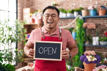 Chinese young man working at florist holding open sign smiling and laughing hard out loud because funny crazy joke.