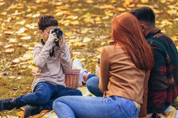Young happy parents having fun with their boy in the park during autumn day. Boy takes a photo of his parents on a picnic