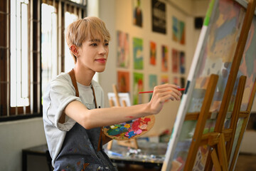 Talented young 20s asian gay man wearing apron painting picture with brush on easel in art studio