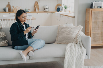 Woman holding banking card, phone making cashless payment sitting on couch at home. Online banking