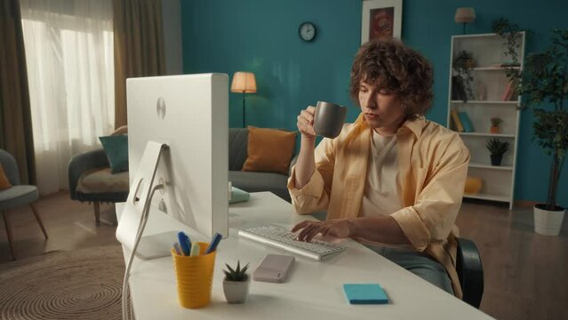 A young curly guy sits at a computer and drinks his morning coffee, tea. Man typing on keyboard, writing email, message, chatting, working online, freelancer. Slow motion.