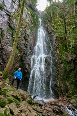 Tourist attraction of Germany - falls of Burgbach Waterfall near Schapbach, Black Forest, Baden-Wurttemberg, Germany. Man hiker in blue jacket standing on stone and looks at flow of falling water