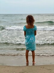 Little girl child in a blue dress stands in the evening on the seashore in cloudy weather rear view, selective focus