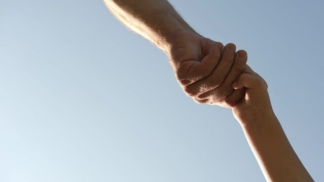 Adult holding a child by the hand on blue sky background. Parent hand reaches out to help child hand in need. Kids Shaking Hands. Children's hand reaches for a daddy. family united. Handshake