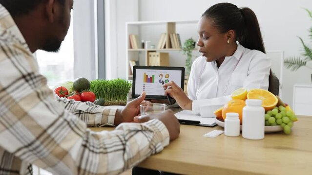 Experienced professional displaying nutrition data graphically via tablet for multicultural man in doctor's workplace. Female expert in diet motivating client to right food choices during treatment.