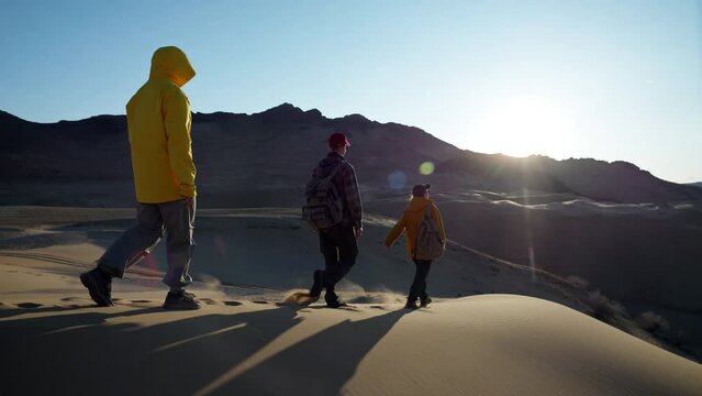 Travel group together goes along sand dune to mountains in sunlight. Wild desert exploration by young hikers in trekking trip, way forward to top