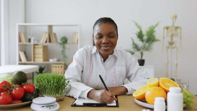 Portrait of african american woman in lab coat holding pen above clipboard while sitting at writing desk in office. Nutrition professional making changes in patient's diet plan in cozy workplace.