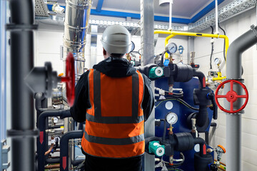 Engineer with back to camera. Man near boiler equipment. Worker in helmet stands in basement. Engineer inspects industrial equipment. Boiler room with pipes. Boiler system installation concept - obrazy, fototapety, plakaty