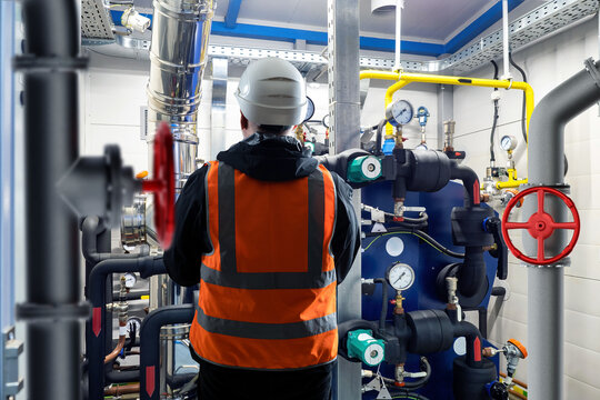 Engineer with back to camera. Man near boiler equipment. Worker in helmet stands in basement. Engineer inspects industrial equipment. Boiler room with pipes. Boiler system installation concept