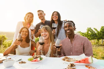 Poster Happy adult friends having fun drinking red wine and eating together with vineyard in background - Multiracial people doing party at summer time in countryside resort - Main focus on center girl face © DisobeyArt