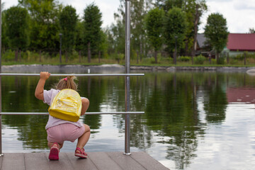 A child girl with a yellow backpack in summer looks at the water in the pond and tries to see the fish there. Leaning over the pere, the child looks at the water in the summer in the park.