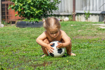 Fototapeta na wymiar Cute funny girl child playing soccer ball in the backyard on a summer sunny day. A child rides a soccer ball on the yard lawn. Girl child plays with a ball on the grass.