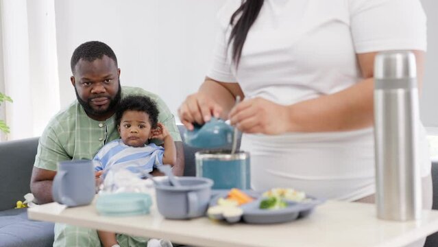 Asian mother prepares food for feeding her 9 months old her cute little baby and African American holds the baby on the sofa. Photo series of family, kids and happy people concept. Parents feed kids.