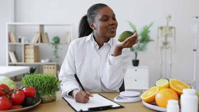 Attractive multiethnic woman in lab coat smiling at avocado on her palm while starting working day at hospital. Cheerful nutrition professional having fun with fruit while describing slimming foods.