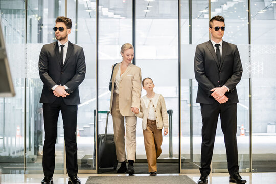 private security concept, two bodyguards in formal wear and sunglasses standing near hotel entrance, cheerful woman and child holding hands and walking with luggage, entering lobby, luxury lifestyle