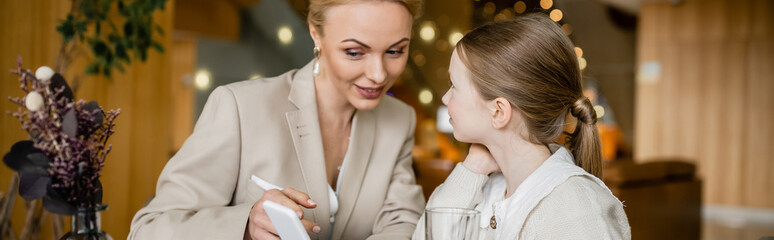 happy mother and daughter spending quality time together, blonde woman holding smartphone near daughter, digital age, working parent and kid, modern parenting, family bonding, banner