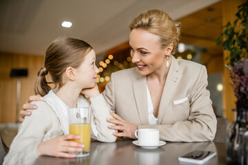 happy mother spending quality time with daughter, blonde woman hugging preteen girl near beverages and smartphone, working parent and kid, modern parenting, family bonding