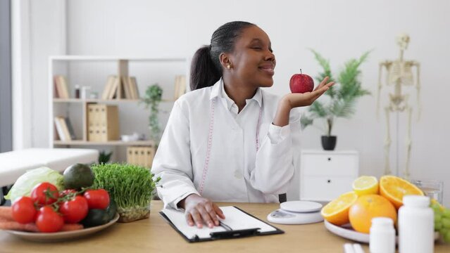 Charming adult lady with measuring tape smelling red apple on palm while noting down nutritional value on paper. Multicultural dietitian in white coat providing with cooking tips with fresh fruits.