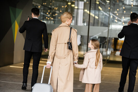 bodyguards walking next to blonde woman and preteen kid, entering hotel, private security, mother holding hands with daughter, people wearing trench coats, safety and protection, back view