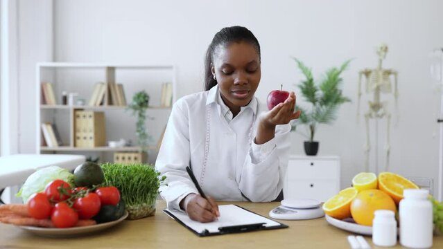 Portrait of smiling female posing with apple close to cheek while doing paperwork in doctor's office. African american nutritionist including client's preferences about fruits into meal plan.