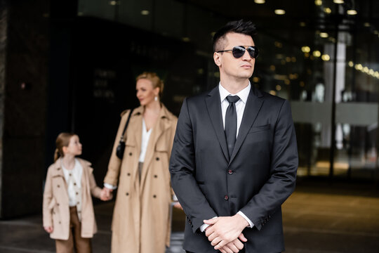 personal security, lifestyle, bodyguard in suit standing near successful woman with preteen child, protecting mother and daughter near hotel, rich life, family travel, private security service