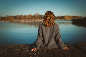 A girl in a coat sits on a wooden bridge in autumn, rear view