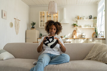 Excited African American woman looking at her first VR headset and feeling excited to get immersed, sitting on sofa in living room. Curious black lady gamer holding futuristic virtual reality glasses