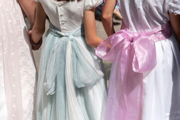 Niñas de primera comunión con elegantes vestidos blancos y lazos de colores caminan cogidas de la mano.