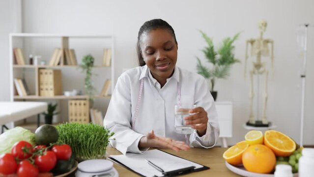 Portrait of multicultural lady posing with glass of clean water while sitting at writing desk in consulting room. Joyful expert in food emphasizing importance of fluid balance on healthy diet.