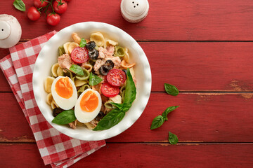 Italian pasta salad. Orecchiette pasta with tuna, tomato cherry, olive, basil and parmesan cheese in plate on red old rustic table background. Traditional Italian cuisine. Flat lay. Copy space.