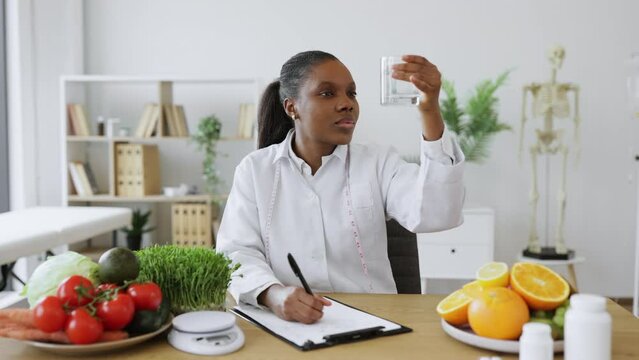 Focused african american woman in white coat checking fluid clearness in glass at doctor's workplace. Effective nutrition specialist advising about drinking water for boosting metabolism at diet.