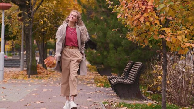 beautiful mature blonde woman with pigtails in trendy beige coat smiles and walks in autumn leaves in the street. Middle-aged woman walks through the city with bouquet of yellow maple leaves